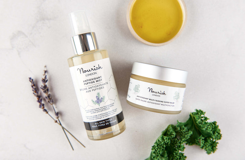 Nourish London Antioxidant Peptide Mist and Antioxidant Multi-Tasking Super Balm displayed with lavender sprigs, kale leaves, and a bowl of golden balm on a light background.