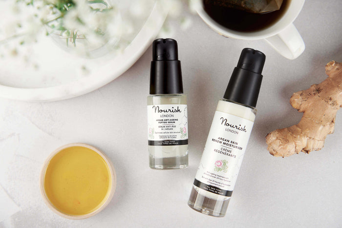 Flat lay of Nourish London skincare featuring Argan Skin Rescue facial oil and Argan Skin Renew Moisturiser, styled with ginger root, herbal tea, and a cup of tea on a light background